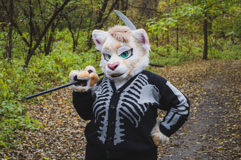 A cougar fursuit looking into the camera on a path in the woods. They are wearing a skeleton cardigan and have a scythe on their shoulder.