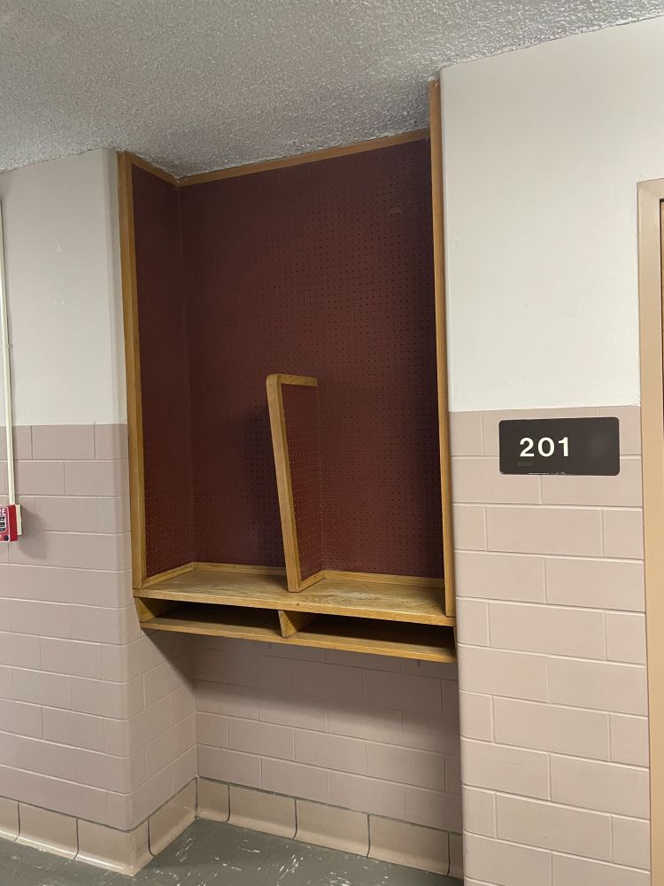 An alcove in a hallway containing a wooden shelf with a vertical divider. The space seems to have once hosted pay phones or other landline phones.