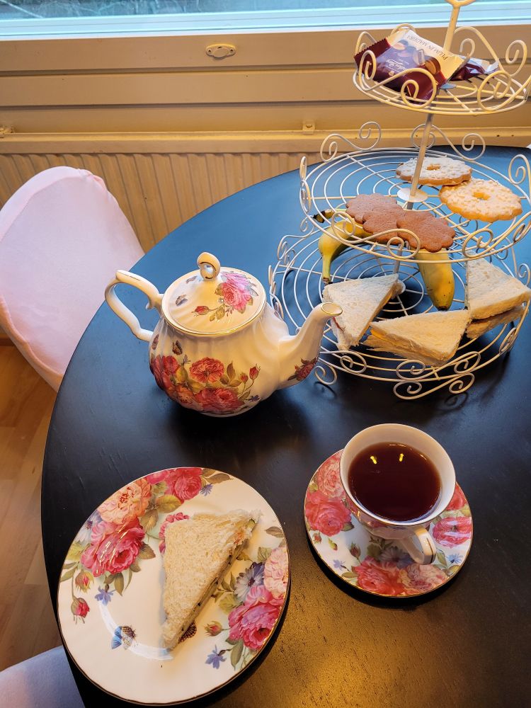 Ruusukuvioinen teeastiasto in action. Kolmikerroksisella tarjottimella on kolmioksi leikattuja leipäsiä, banaania, piparkakkua ja pikkuleipää sekä plums in madeira- konvehteja