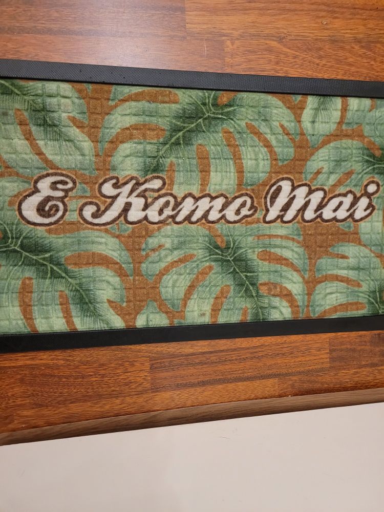Kynnysmatto, jossa peikonlehtikuvio ja suurilla ystävällisillä kirjaimilla teksti E Komo Mai, eli tervetuloa havaijiksi