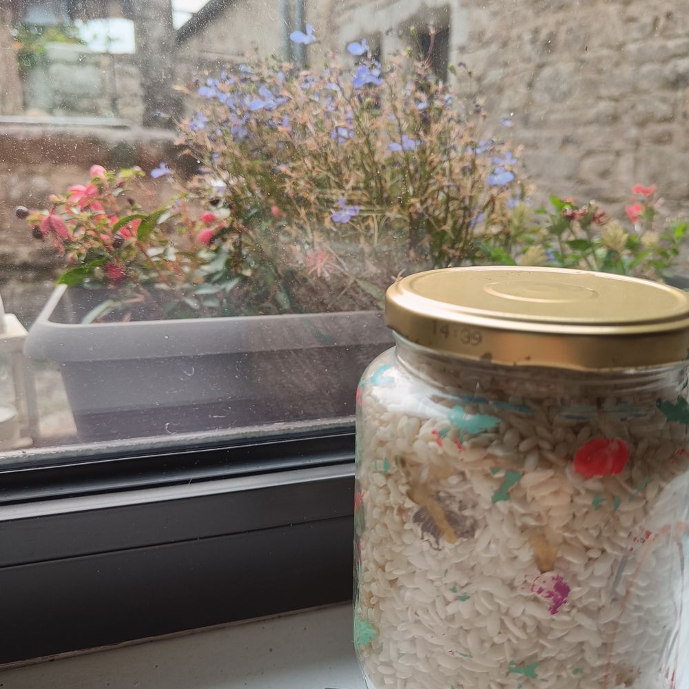 Ein dekoriertes Glasgefäß (die Farben sind verblasst, es ist Reis darin) mit Blumen hinter dem Fenster.