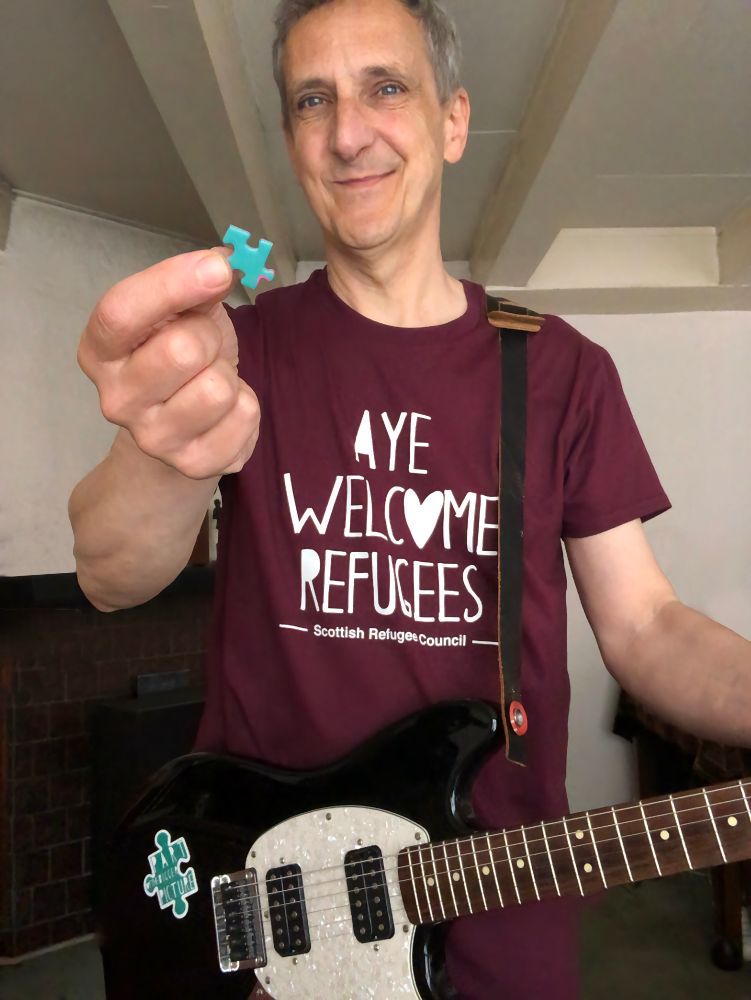 Old guy standing looking at the camera, wearing a maroon t-shirt with the text AYE WELCOME REFUGEES on it. A black and white electric guitar (with a green and white sticker with text that reads PART OF THE BIGGER PICTURE) is hanging around his shoulders. In his right hand he is holding a green puzzle piece.