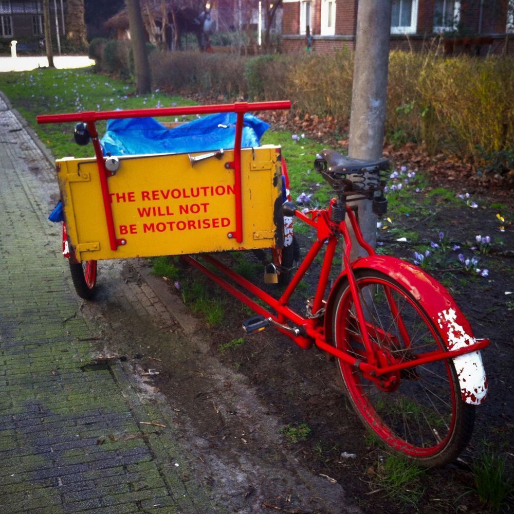 Red bike parked at the side of a foot path. At the front of the bike is yellow container with the words "THE REVOLUTION WILL NOT BE MOTORISED" on it