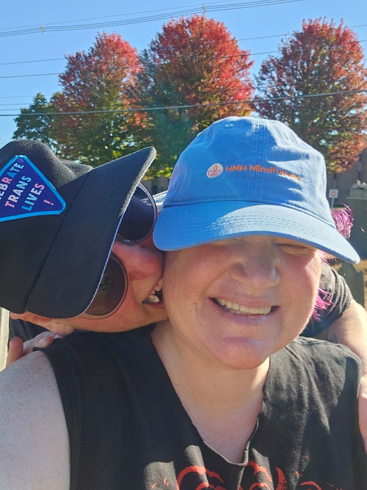 A photo of two white nonbinary people. I'm smiling, wearing a blue ball cap and black tank top. Liz is biting at my neck from behind, wearing a black ball cap and sunglasses.