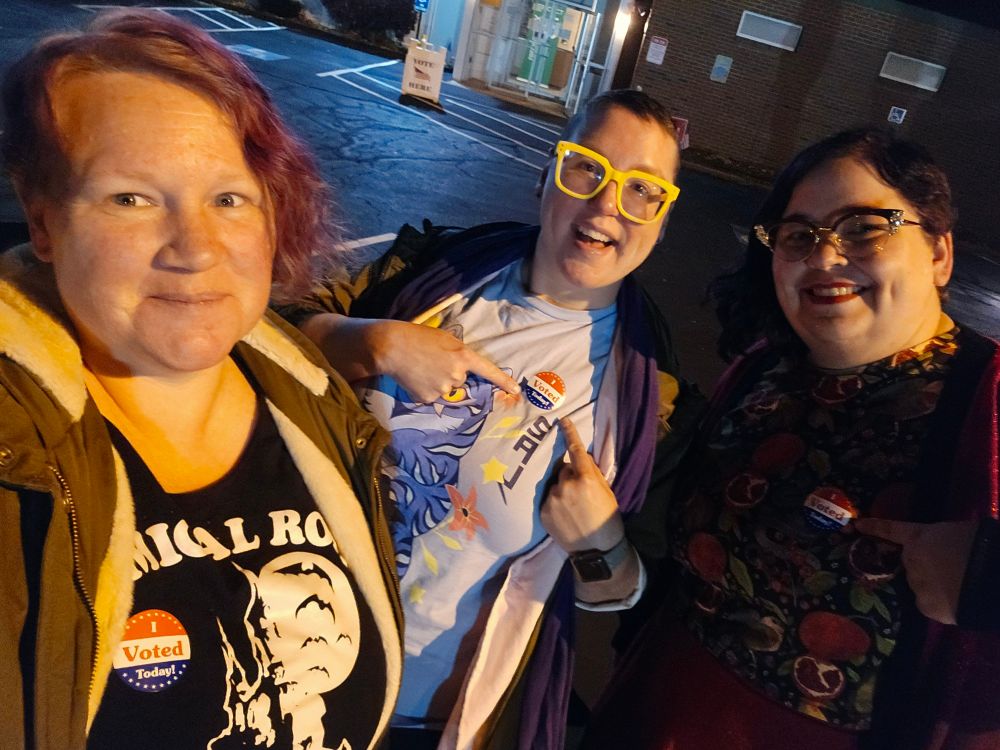 A photo of three white people standing in a parking lot in t-shirts and jackets. They all have I Voted Today! stickers and are smiling at the camera.