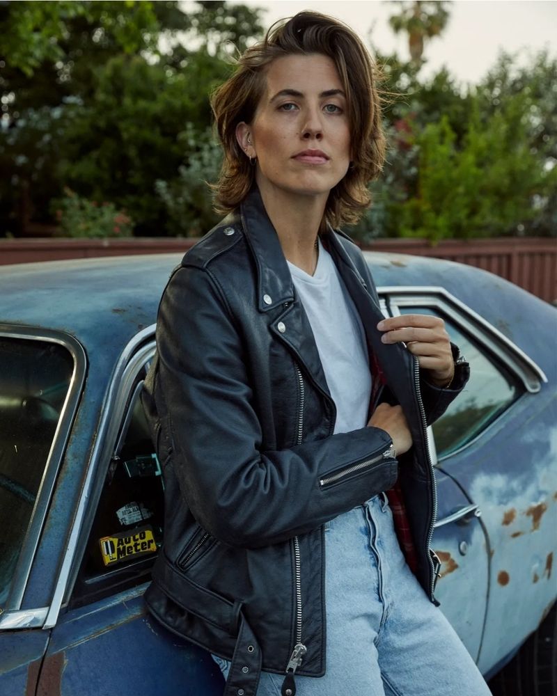 A white non-binary person with toussled dark hair leaning against a car and wearing a white t-shirt, leather jacket, and jeans.