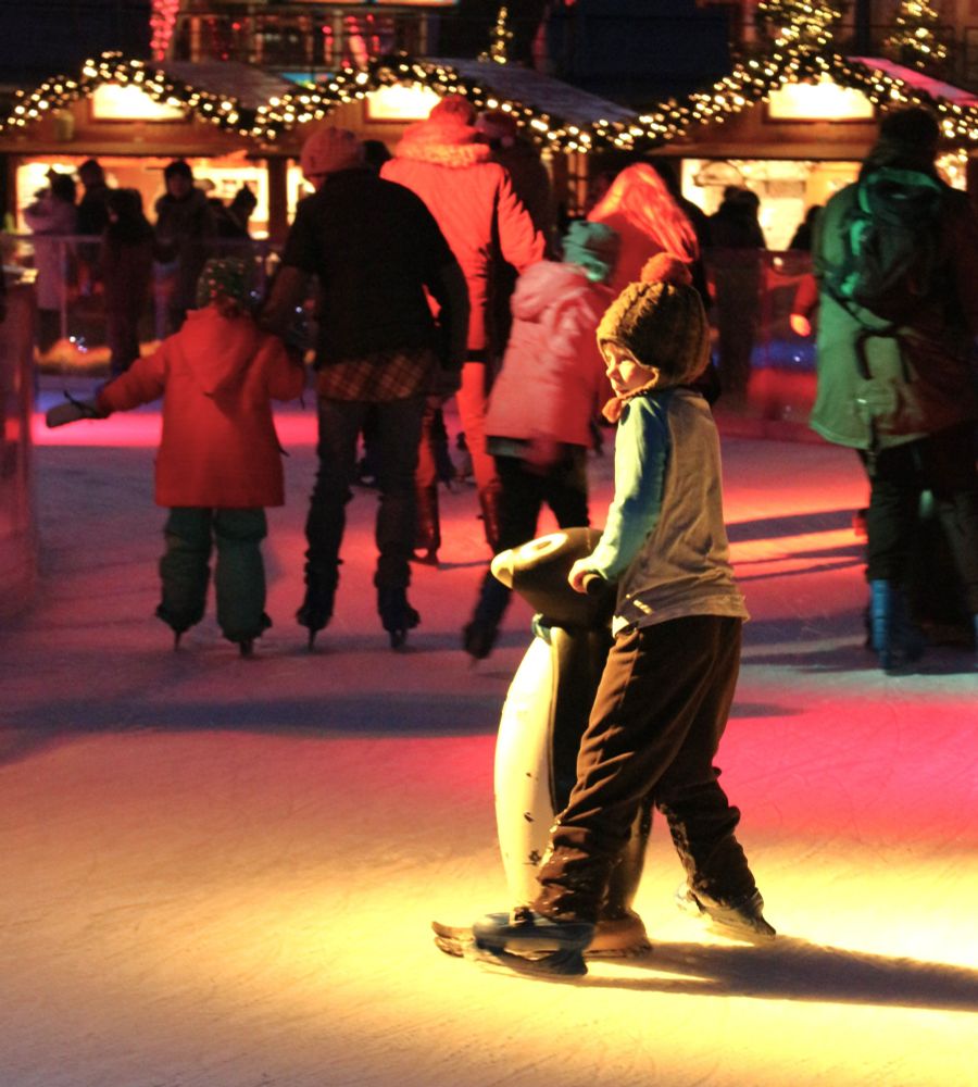 Spot lit young skater sideways on, wearing a woolly hat with face looking to left of frame. with other skaters and cabin shops behind. Lots of reds, pinks and yellows.