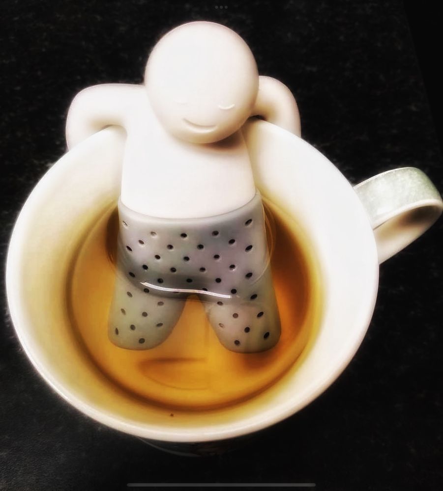 View of tea cup from above with relaxed man tea strainer, hooked on sad and legs in the yellowing liquid. 