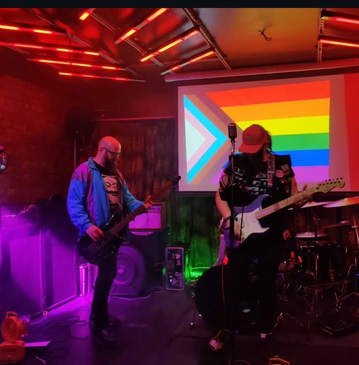 The digital post-rock duo, West Echo Three, onstage in front of a projection of the progress pride flag. 