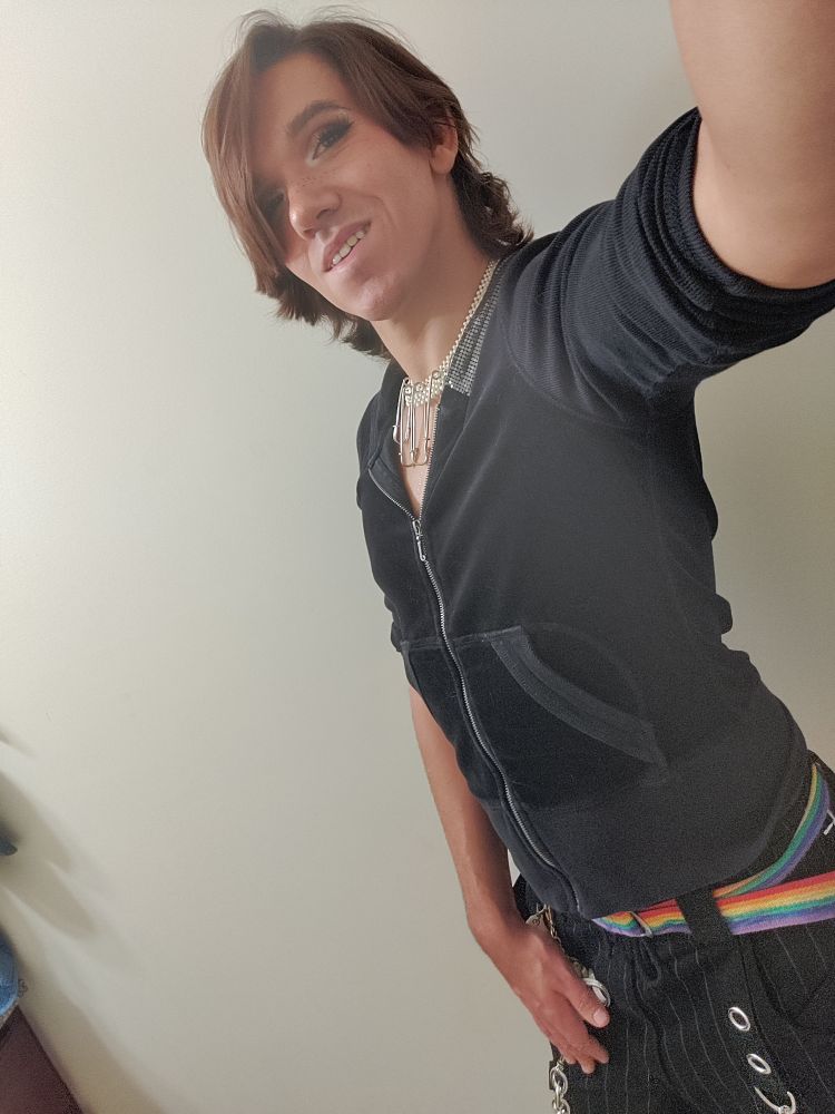 A non binary person wears a black Juicy Couture hoodie and black pinstripe Tripp pants with a rainbow belt. A pearl necklace with large safety pins hangs on their neck. They smile into the camera. 