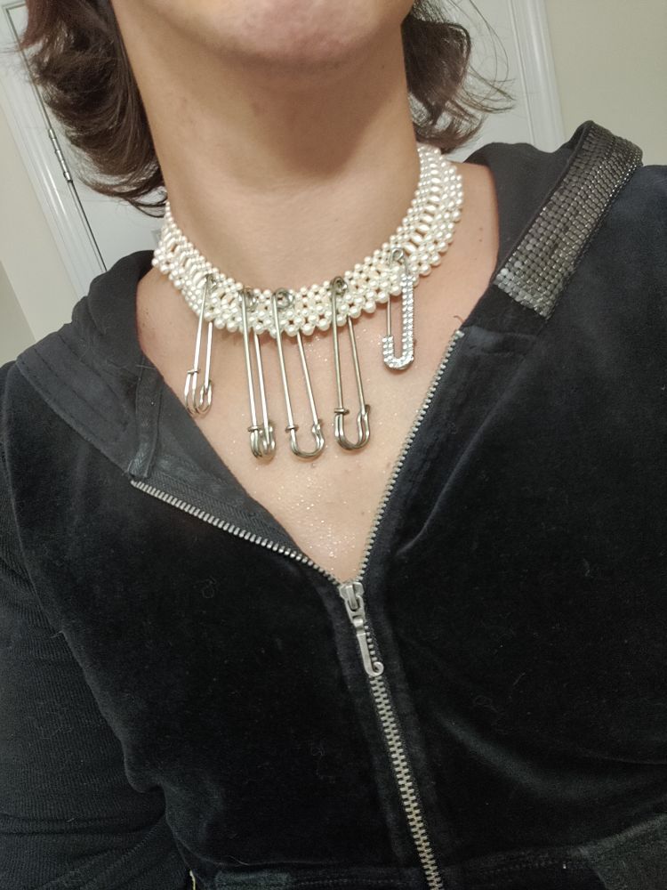 A closeup of a person wearing a pearl necklac with four large safety pins handing from it, one of which is bejeweled with a "J". The person also wears a black velour Juicy Couture hoodie with onyx mesh panels lining the hood and a silver "J" pull on the zipper. 