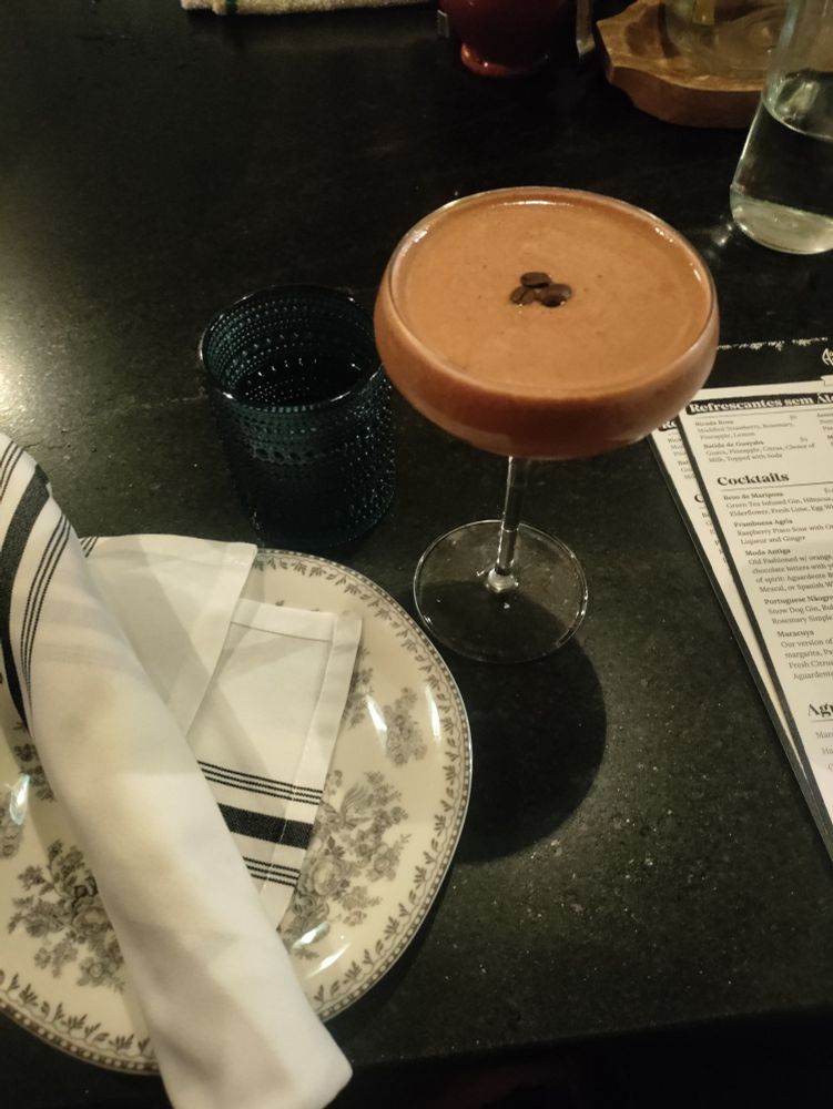 A place setting at a bar with an ornate plate, cloth napkin, a blue glass of water and a dark brown espresso martini. 