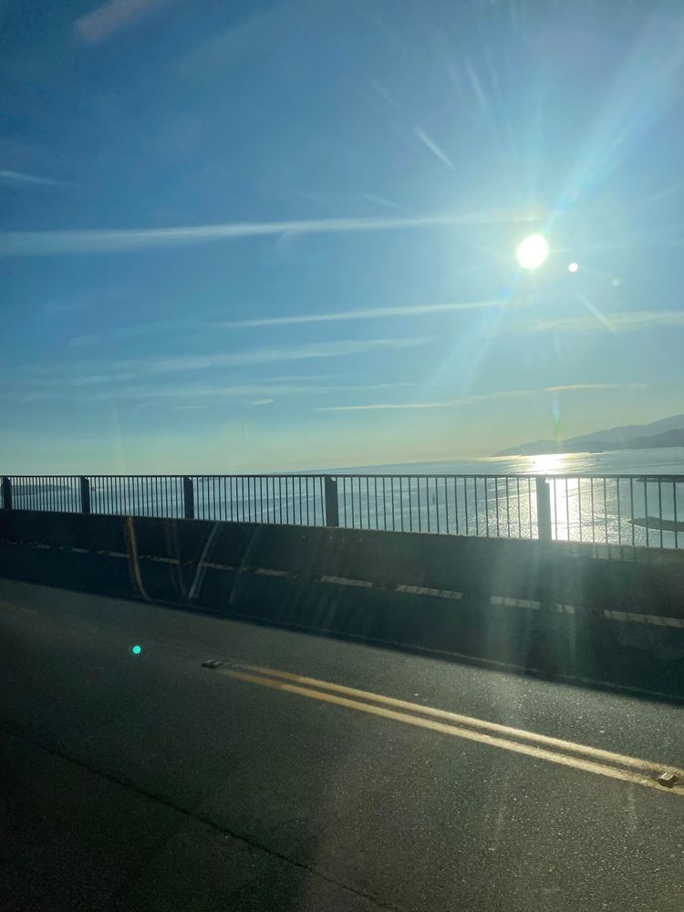 the sun is low in the mostly blue but whispy sky over the water as seen from the window of a bus going over lions gate bridge