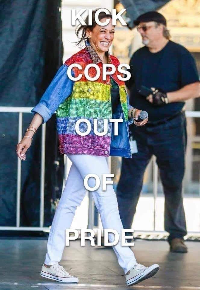 The image shows a Kamala Harris wearing a colorful rainbow-patterned jacket, a black top, white pants, and white sneakers. They are holding a microphone and smiling. A person in the background is wearing a black shirt, cap, and glasses. The text overlaid on the image reads "KICK COPS OUT OF PRIDE." The scene suggests a public or outdoor event, possibly related to LGBTQ+ pride.