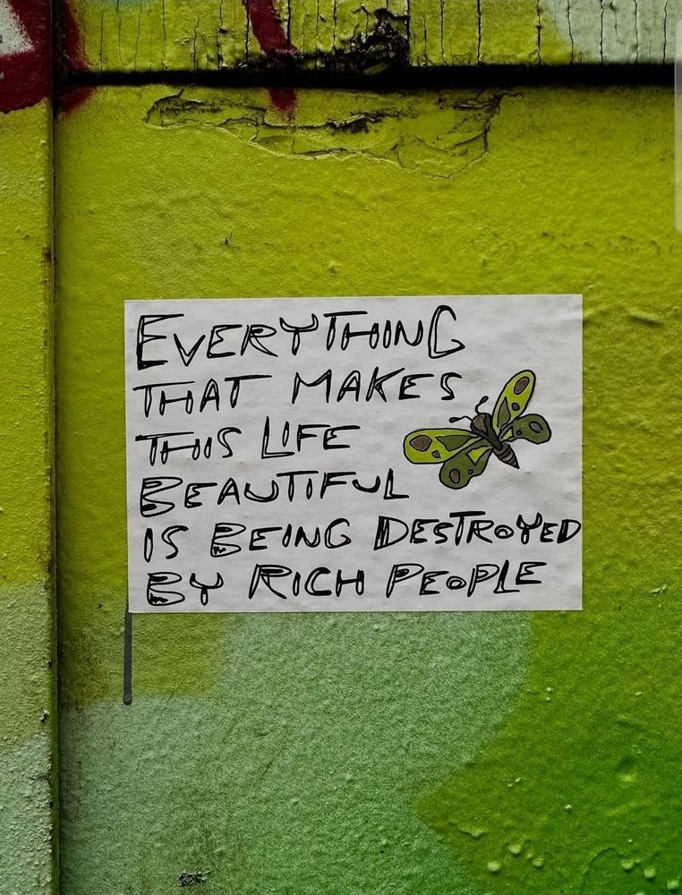 The image shows a piece of graffiti art on a textured wall painted in various shades of green. The wall has visible cracks and weathering, adding to the urban aesthetic. 

In the center, there's a white paper or poster with handwritten text in bold, irregular black letters. The text reads: "Everything that makes this life beautiful is being destroyed by rich people." 
