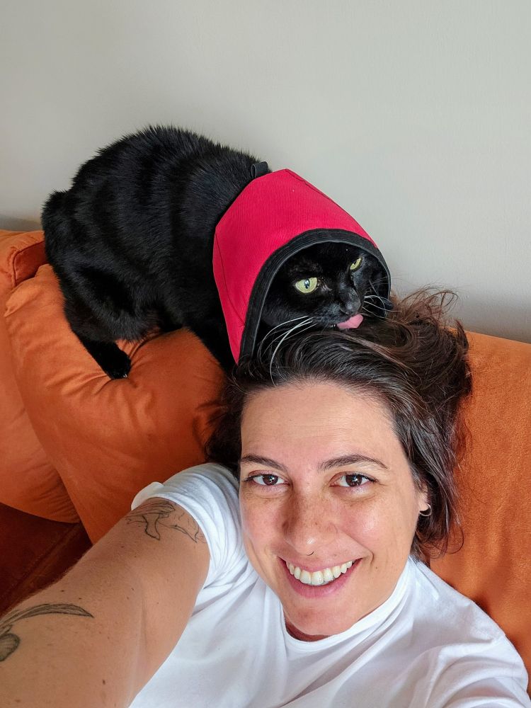 Selfie em que tô encostada num sofá sorrindo e em cima do encosto tá a Cleo lambendo meu cabelo. Cleo é uma gata preta que tá usando um colar elisabetano vermelho. Eu sou uma mulher branca de olhos e cabelos castanhos.