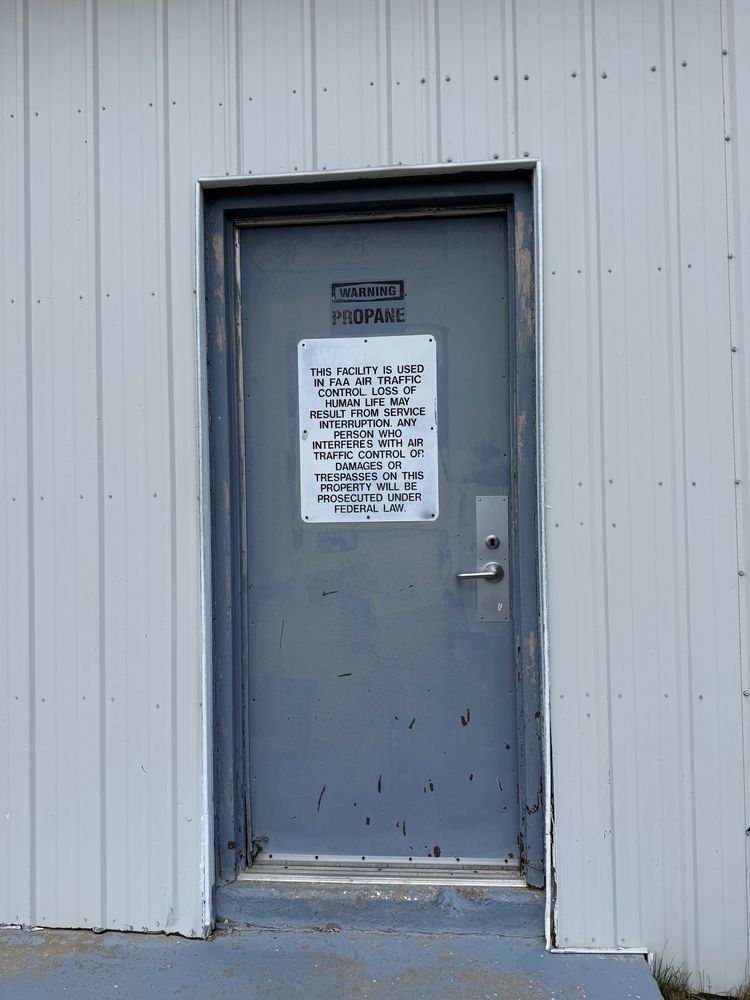 Door of the building in the mid ground. It reads:

WARNING PROPANE
THIS FACILITY IS USED IN FAA AIR TRAFFIC CONTROL. LOSS OF HUMAN LIFE MAY RESULT FROM SERVICE INTERRUPTION. ANY PERSON WHO INTERFERES WITH AIR TRAFFIC CONTROL OP.
DAMAGES OR
TRESPASSES ON THIS PROPERTY WILL BE PROSECUTED UNDER
FEDERAL LAW.