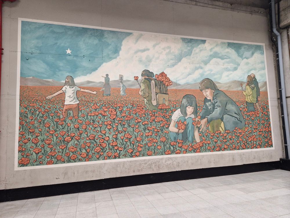 Des femmes et des enfants parcourent le désert fleuri chilien en ramassant des fleurs. Une étoile blanche pointe à l'horizon, au milieu d'un ciel bleu. Une femme porte à sa veste l'insigne des associations de recherche des détenus disparus avec le slogan "Dónde están?" et la photo du portrait d'une femme et d'un homme en noir et blanc. 