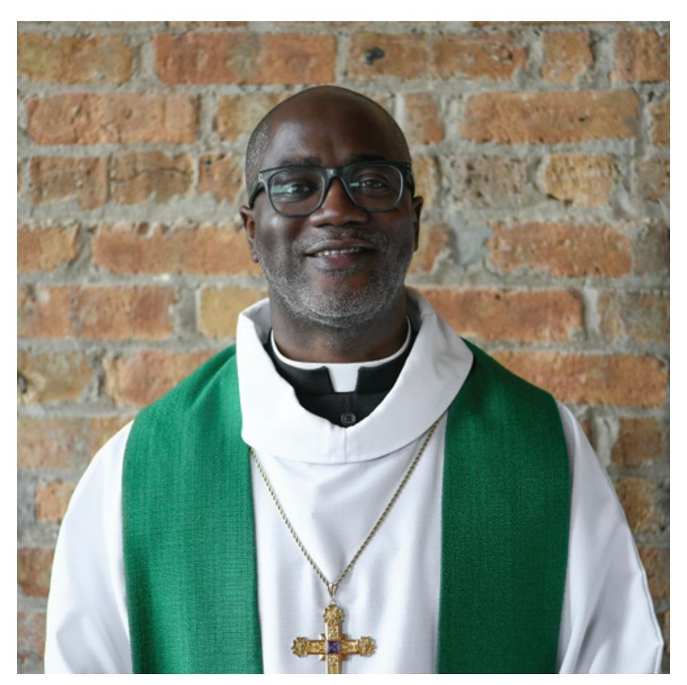 A photo of Yehiel Curry, a black African American man wearing glasses. He is bald. He is smiling slightly. He is dressed in clerical garb, including a large gold cross.