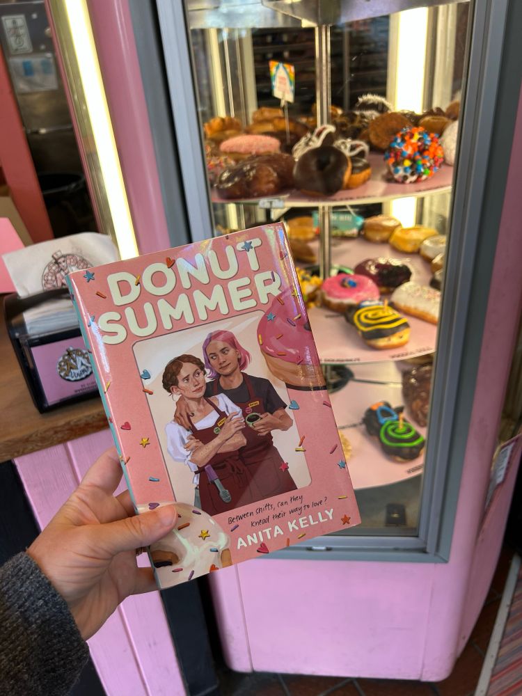 My hand holding DONUT SUMMER by a rack of donuts inside Voodoo Doughnuts