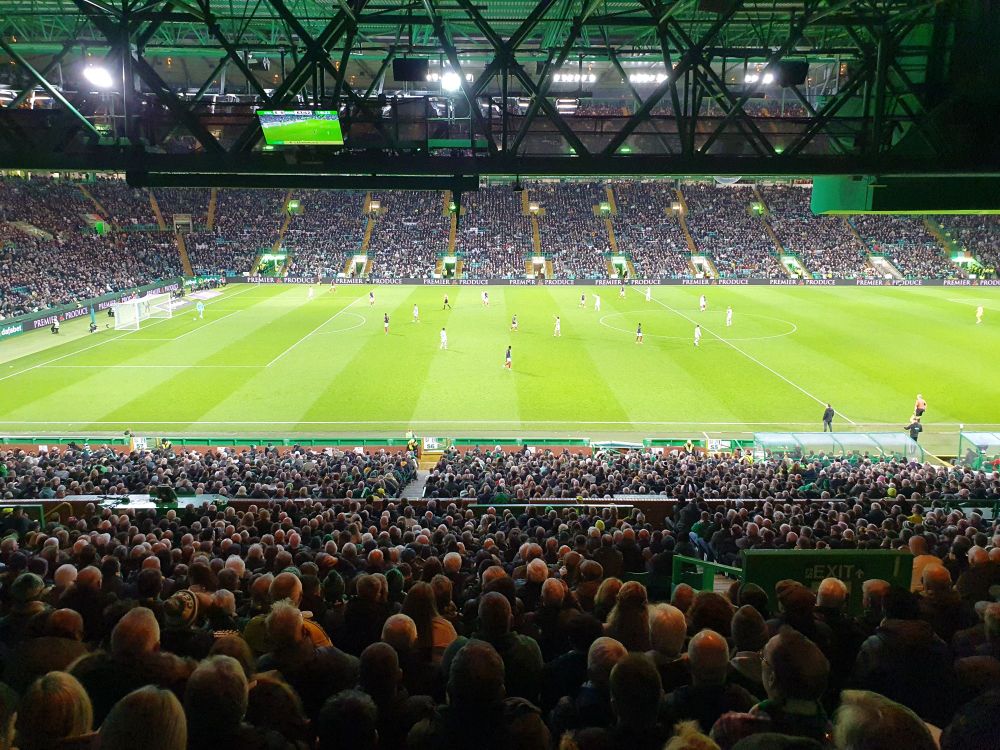 View of a football match in progress under floodlights at Celtic Park. Looking over heads of a packed crowd from the back of the main stand.