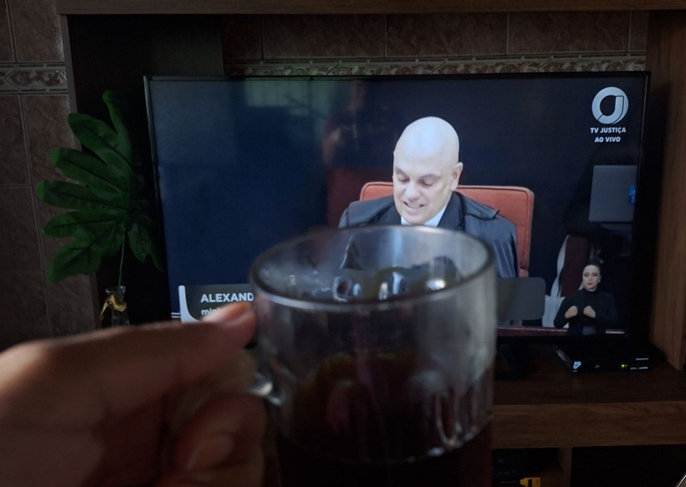 Imagem de uma TV ligada na TV Justiça com a imagem do ministro Alexandre de Moraes. Em primeiro plano, uma xícara transparente com café 