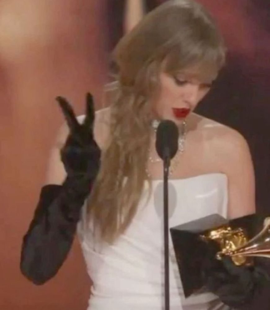 Taylor Swift at the Grammy’s in a white dress and black gloves holding up two fingers 
