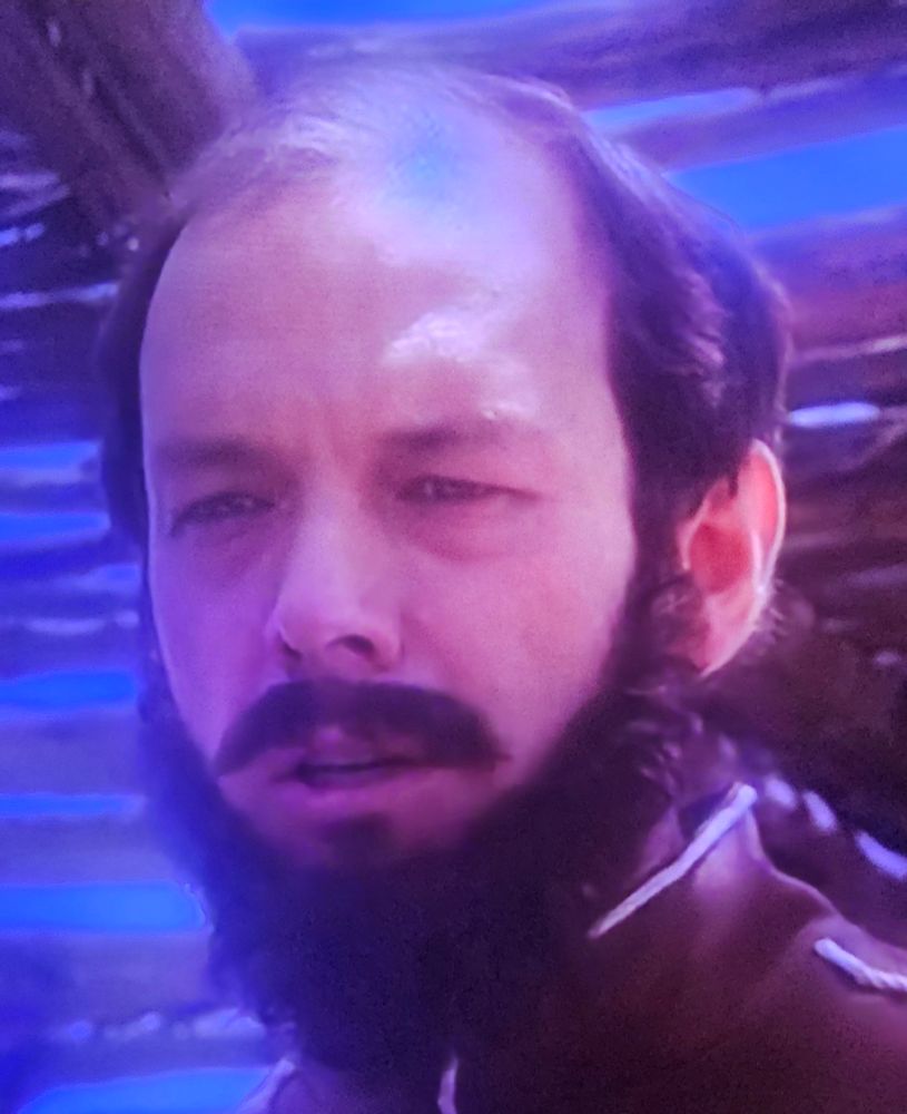 Nahaufname aus einem Film. Mann dunklem Haar und merkwürdig aussehendem Barr.