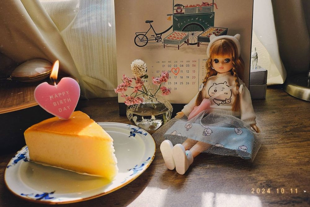 Una muñeca Licca vestida con atuendo de Chiikawa al lado de un trozo de pastel con una vela que dice "Happy Birthday" y unas flores en un florero pequeño.
Detrás hay un calendario con la fecha 10 de octubre marcada con un corazón