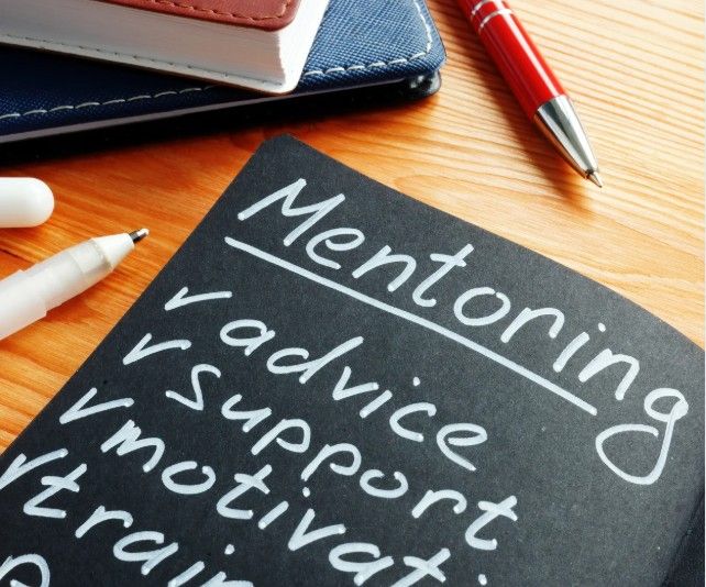Close-up of a piece of paper labeled 'Mentoring' with words such as 'advice,' 'support,' and 'motivate' written on it, alongside a red pen, a white marker, and a notebook on a wooden desk.