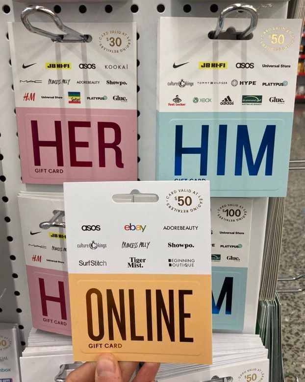 Gift cards on display in a store. One says “her” in pink, and another says “him” in blue. Between them, a hand is holding up an orange gift card which reads “online.”