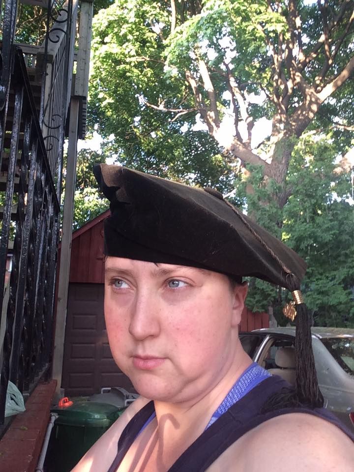 A photo of a noticeably younger Emily, standing outdoors, looking off to the left mock-seriously, wearing a graduation cap. If you look closely, you can see that it's a cheap velvet octogonal doctoral cap. She is wearing a black tanktop with visible bra straps. 