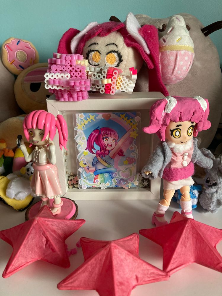 A "shrine" for my UTAU, Yuri Tamaki. On the left is a figurine holding a microphone. On the right is a ball jointed figure. In the center is a Yuri plushie, a framed and decorated image of Yuri in a rainbow outfit, and a Perler bead figure lying on its back. In the very front are 3 paper stars. On the edges are 6 Pokémon plushies, much smaller than the rest of the objects.