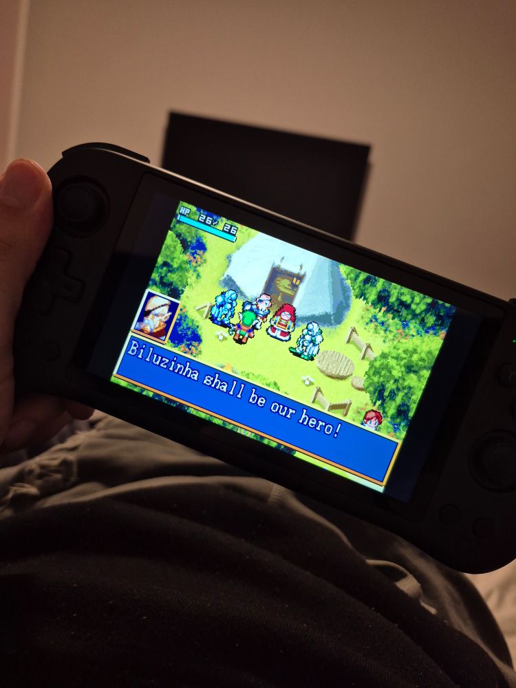 Foto de um console handheld com um jogo em pixel art de personagens reunidos do lado de fora de uma barraca e uma caixa de texto dizendo "biluzinha shall be our hero"