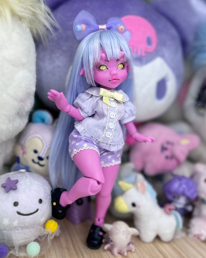 a small pink demon doll in pastels posed with plushies.