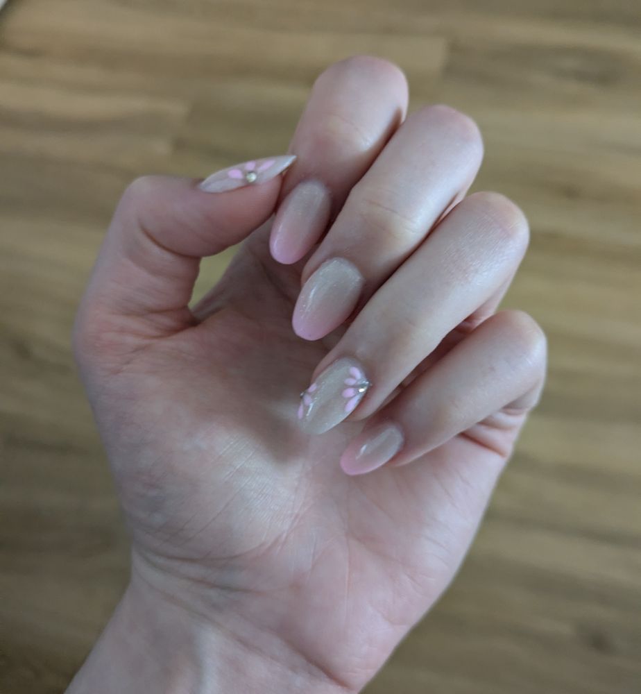 Photo of a hand with a new set of gel nails. The thumb and ring finger have pink flowers with rhinestones. The pointing finger middle finger and the pinky have a pink to light beige ombre