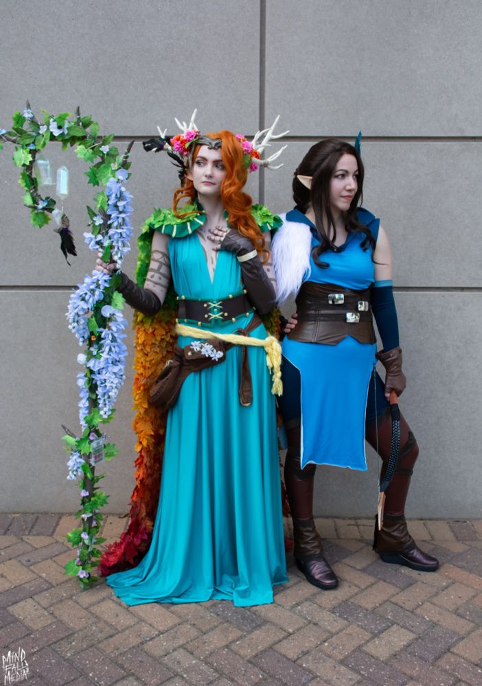 Clya, dressed as lvl 20 Keyleth, stands on the left of the image looking off to the side. Besides her stands @dragonsinparis, dressed as TLOVM Vex, who looks over her side. They stand in front of a basic stone background. 