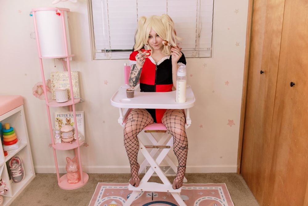 Medusa dressed up as Harley Quinn and sat in the nursery's high chair, There's a bottle of milk on the tray as well as a puddin' cup. She's holding a red pacifier with a dollop of pudding on it, and a pudding covered spoon. 