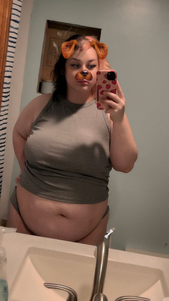 globbie stands at their bathroom sink taking a mirror selfie with their phone. they're wearing a grey high necked sleeveless shirt and grey panties. their shirt is cropped, showing off their soft squishy tummy. globbie's boobs look huge in this shirt, and their thick curvy body is accentuated by the cut and drape of the fabric. globbie's black and faded pink split dyed hair is brushed out and fluffy, matching the cute dog snapchat filter they have on the photo. the filter gives them rusty brown color short, floppy dog ears and a dog nose/face. there's also sharp black cat eye eyeliner as part of the filter. 