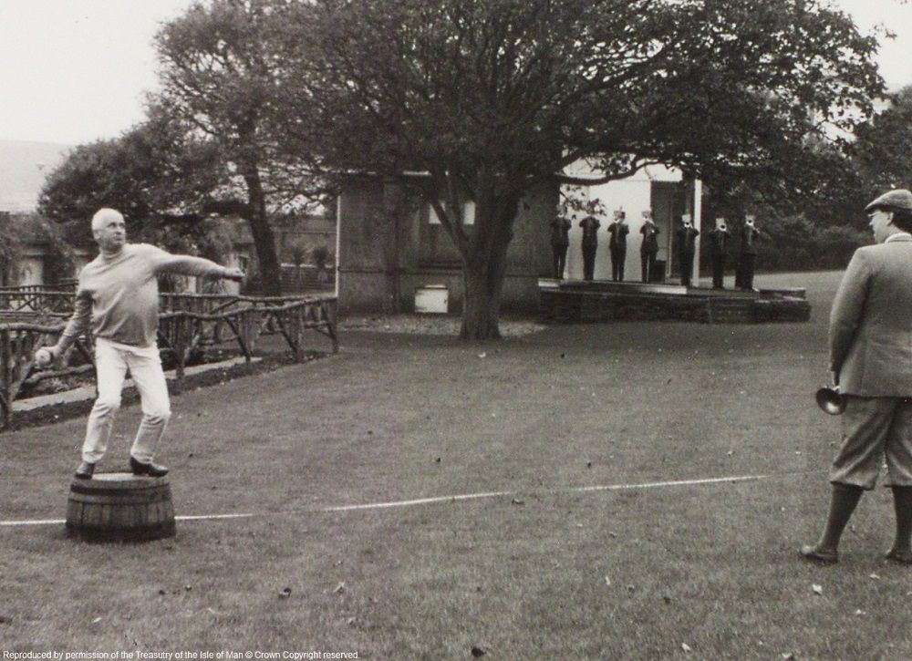 Black and white photograph (dating circa 1980s) of a haggis-hurling contestant in Villa Marina Gardens. He is standing on a whisky barrel and is poised to hurl the haggis. There is a tape measure laid out on the ground behind him. A man in plus fours and a cap looks on. In the background, under a tree, there is a line of trumpeters.  