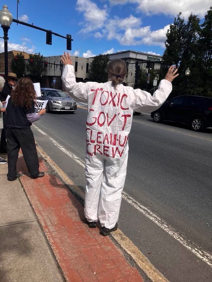 A middle aged woman wearing what looks like a lab or hazmat jumpsuit with the words TOXIC GOVT CLEANUP CREW written on the back in red and black marker. 