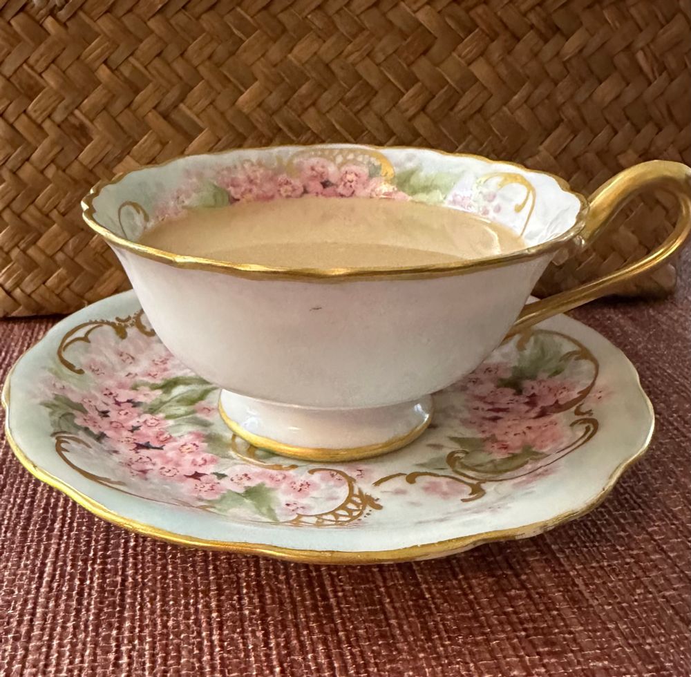 A T&V Limoges tea cup and saucer with pink flowers, green leaves, and gilt curlicues and trim. The background is a watercolor-esque white, pale pink, and pale blue.