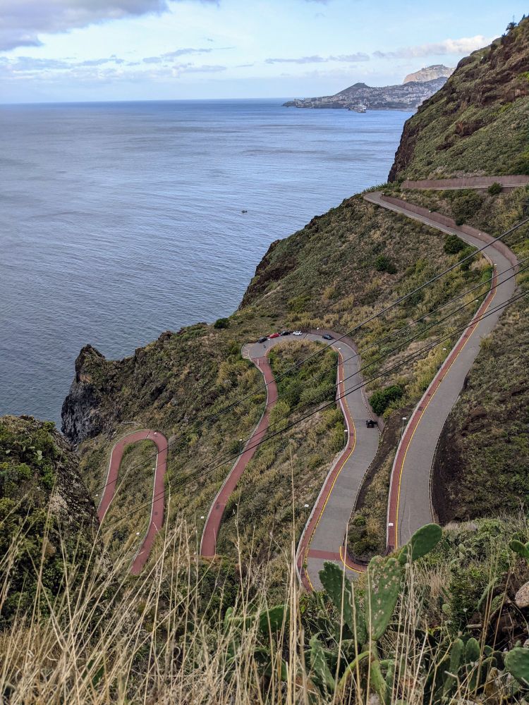 Canico, Madeira. Von rechts oben im Bild führt eine Serpentinenstraße nach links unten durch grün-gelbe Vegetation, im
Hintergrund blau-graues Meer. Im oberen Teil ist die Straße grau mit einem roten Fußgänger-/Fahrradstreifen, im unteren für KfZ gesperrten Teil ausschließlich rot.
