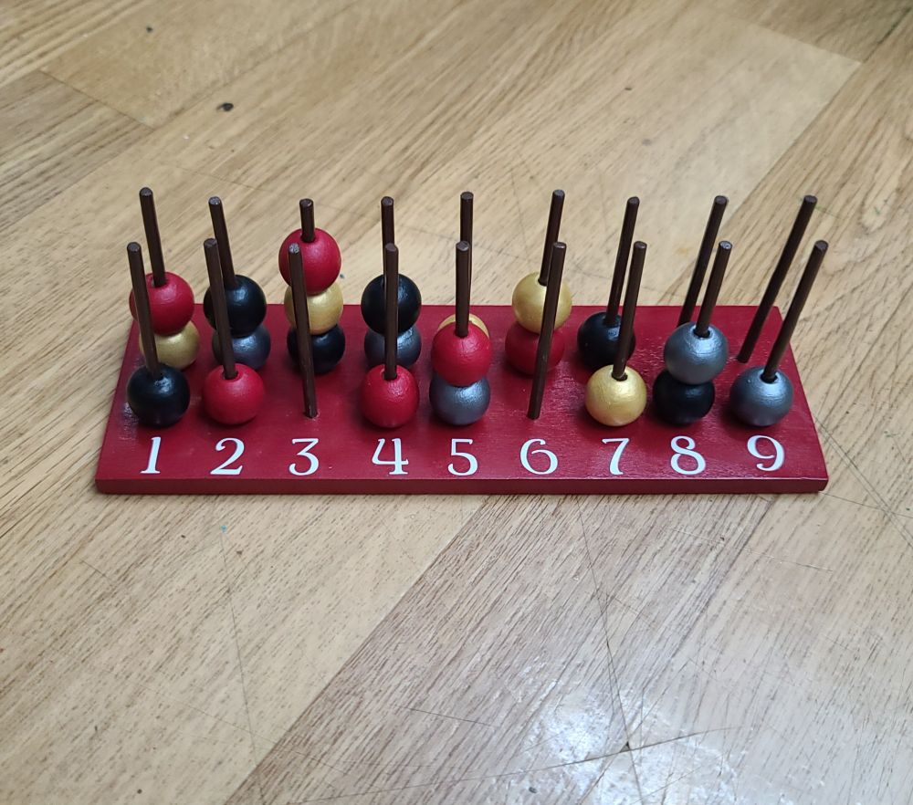 a wood spell slot tracker. the base is red with white numbers. the pegs are black. it has red, gold, black, and silver beads.