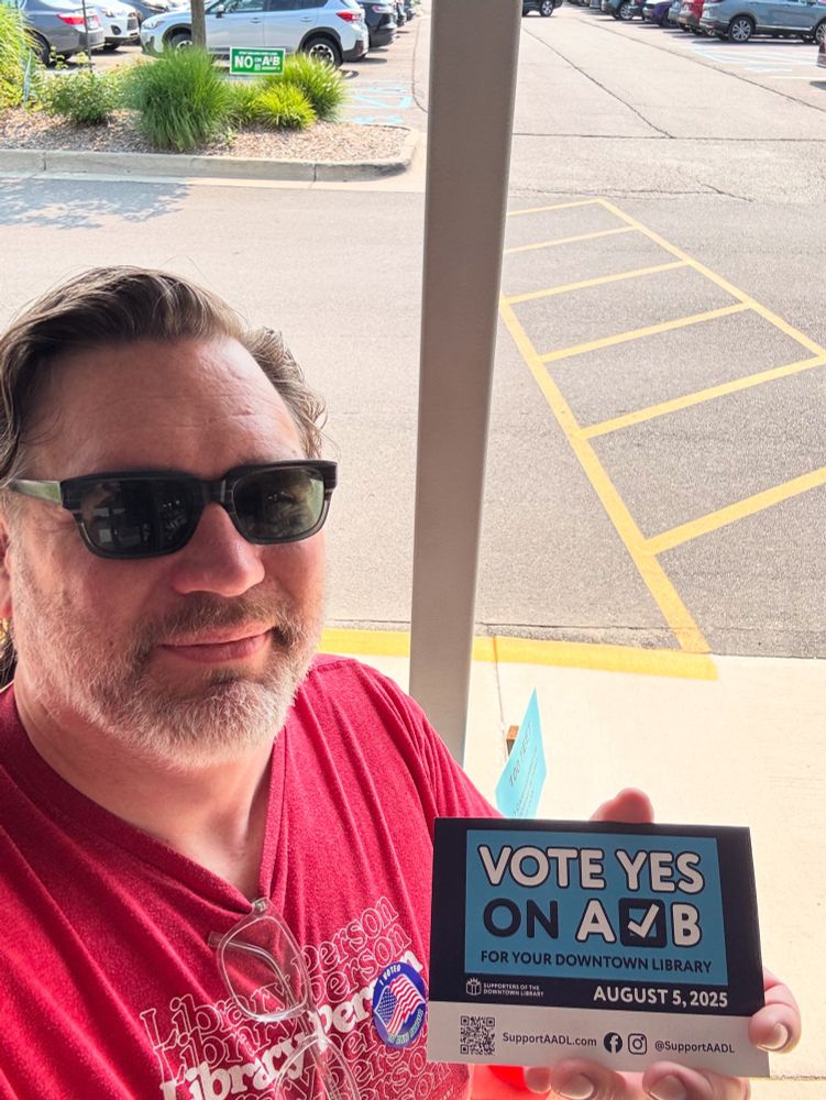 Man in sunglasses wearing a “Library Person” T-shirt holding a “vote yes on a & b” postcard 