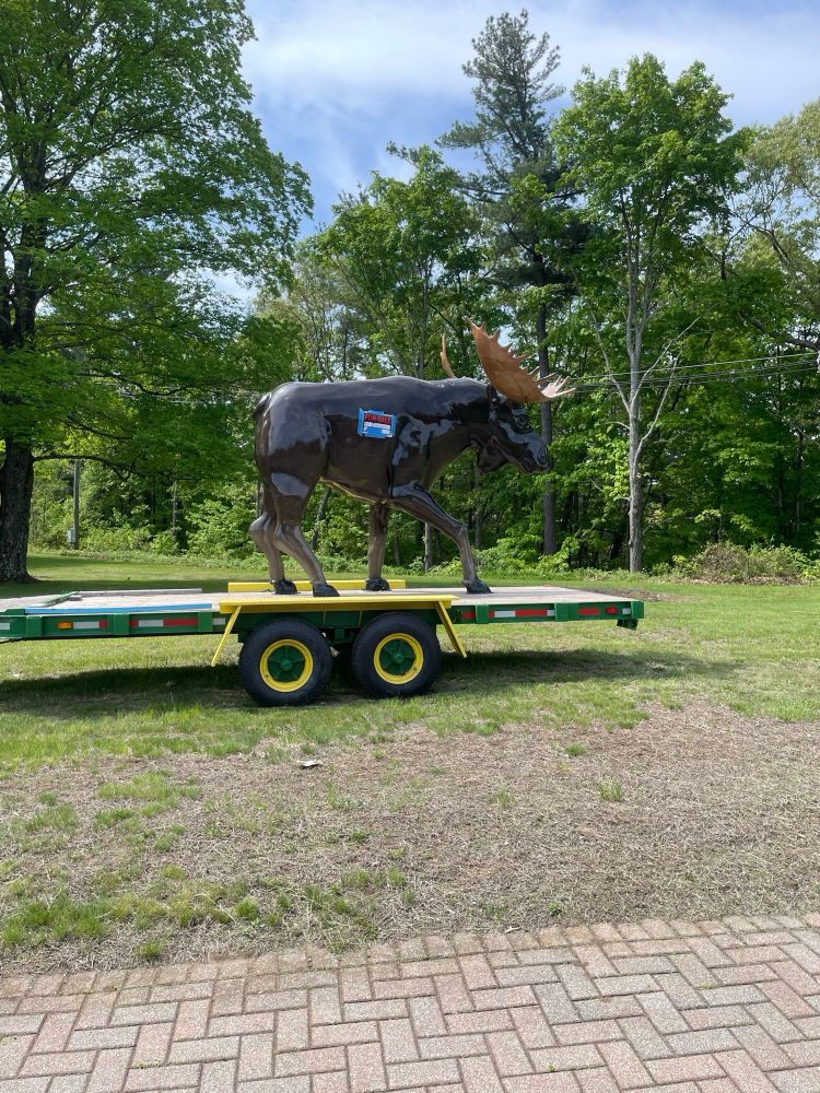 A huge fiberglass moose sculpture with a for sale sign on him, standing proudly on a flatbed truck by the side of the road.