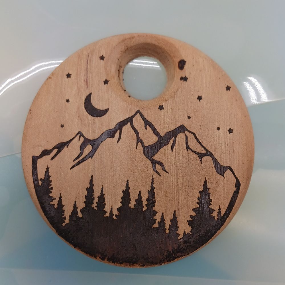 A round wood pendant with a burned in image of a pine tree and mountain scene. 