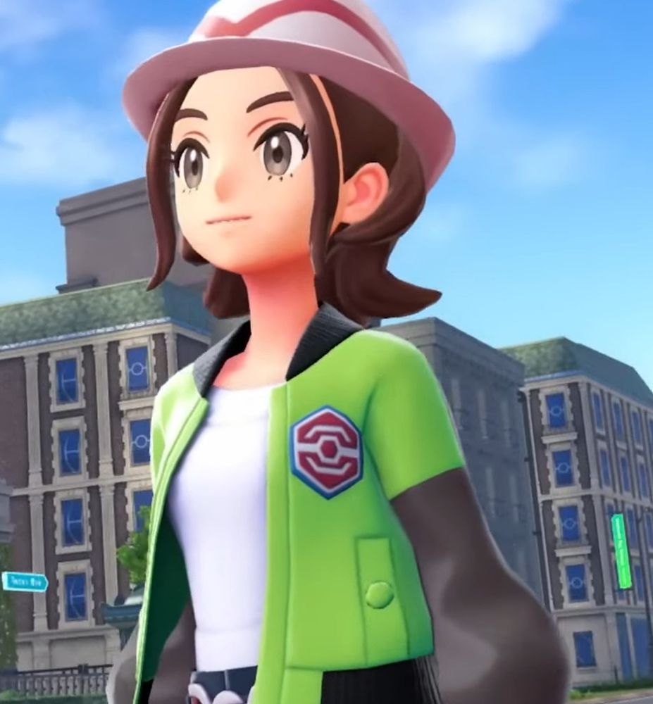 A woman in a black black and green varsity jacket, with brown hair, and a white fedora. 