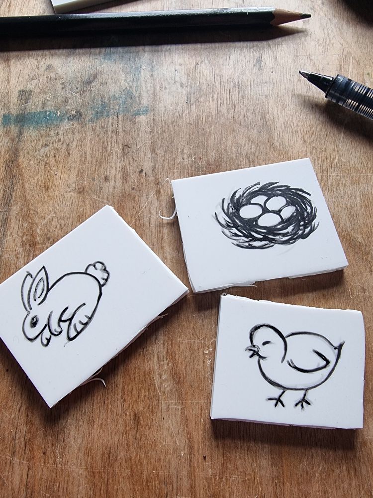 3 small white stamp blocks on a wooden, ink-stained table. Each has a simple pen drawing of a nest with eggs, a rabbit, and a chick, respectively 
