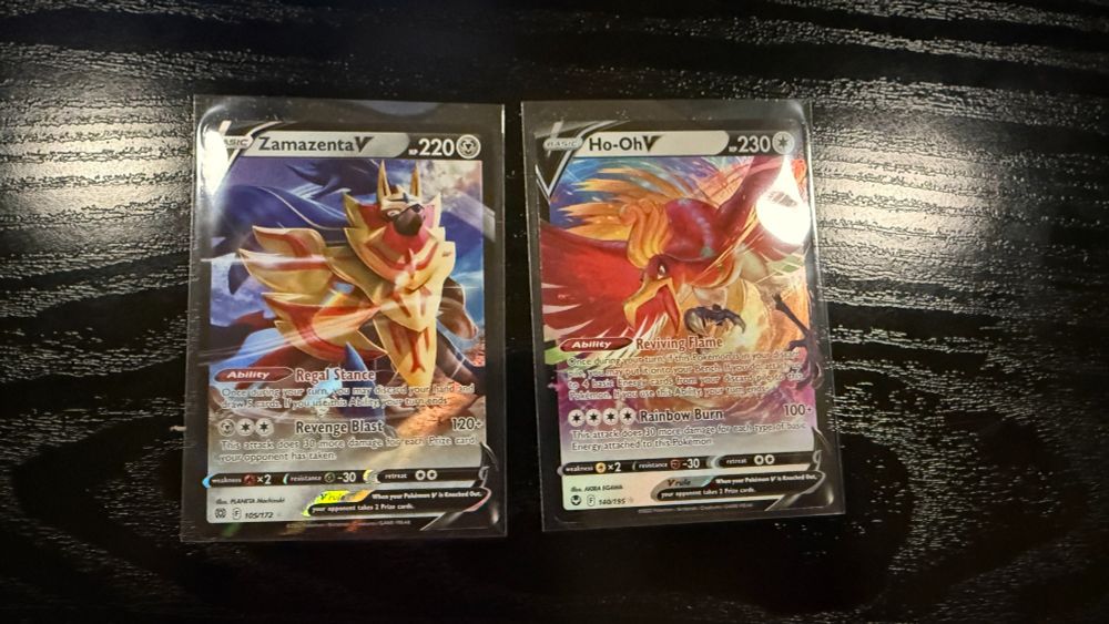 Close-up of two Pokémon TCG cards displayed in protective sleeves on a dark wooden surface. On the left is Zamazenta V, featuring artwork of the legendary shield Pokémon with a detailed background. On the right is Ho-Oh V, showcasing the vibrant and majestic firebird Pokémon surrounded by fiery hues. Both cards highlight their abilities and moves with crisp text.
